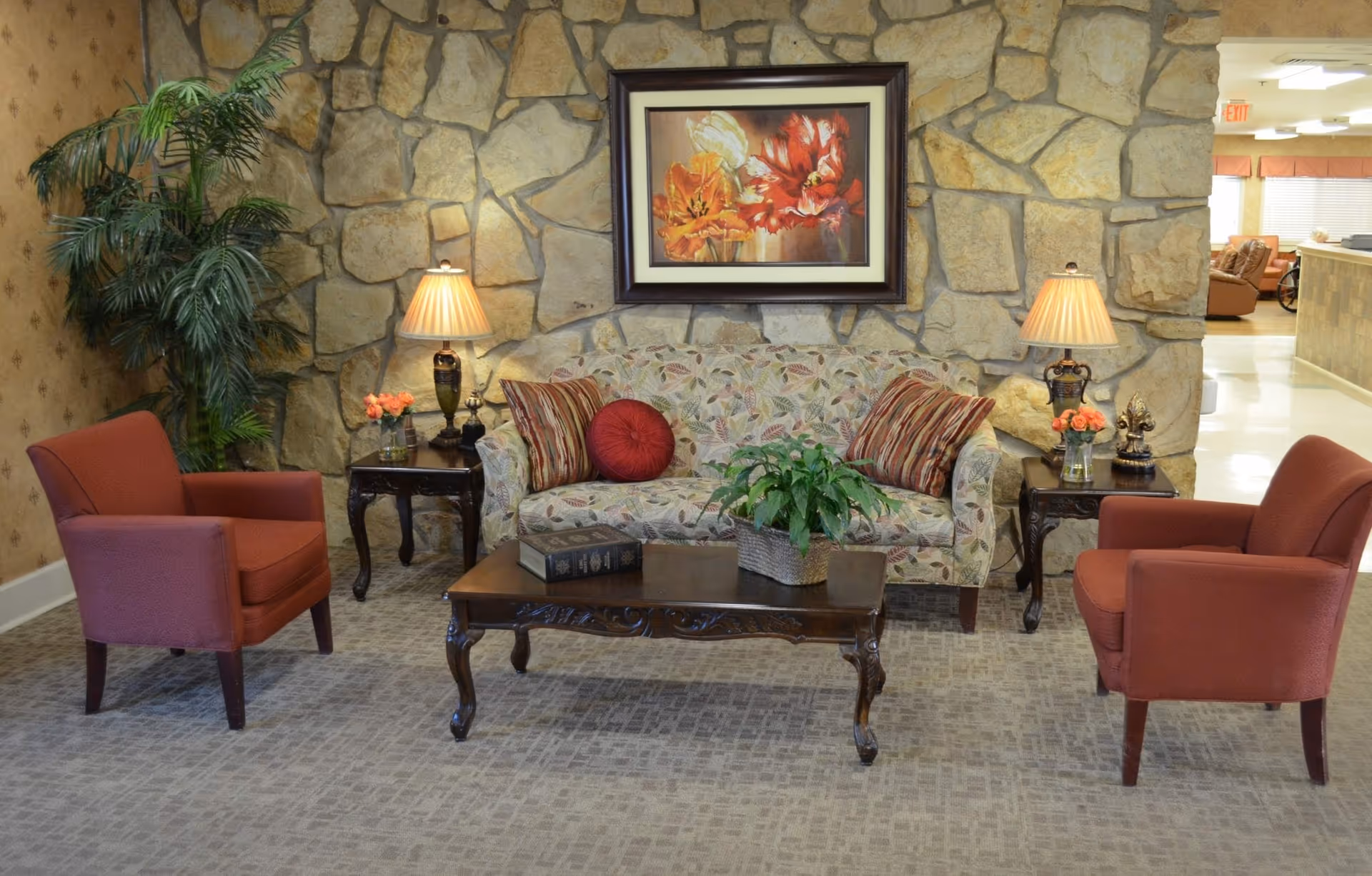 A cozy seating area with a floral sofa, two red armchairs, a wooden coffee table, table lamps, and a stone accent wall with framed artwork.