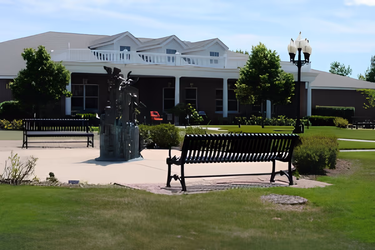 Outdoor courtyard with benches, a small fountain sculpture and lawn in front of a senior living building.