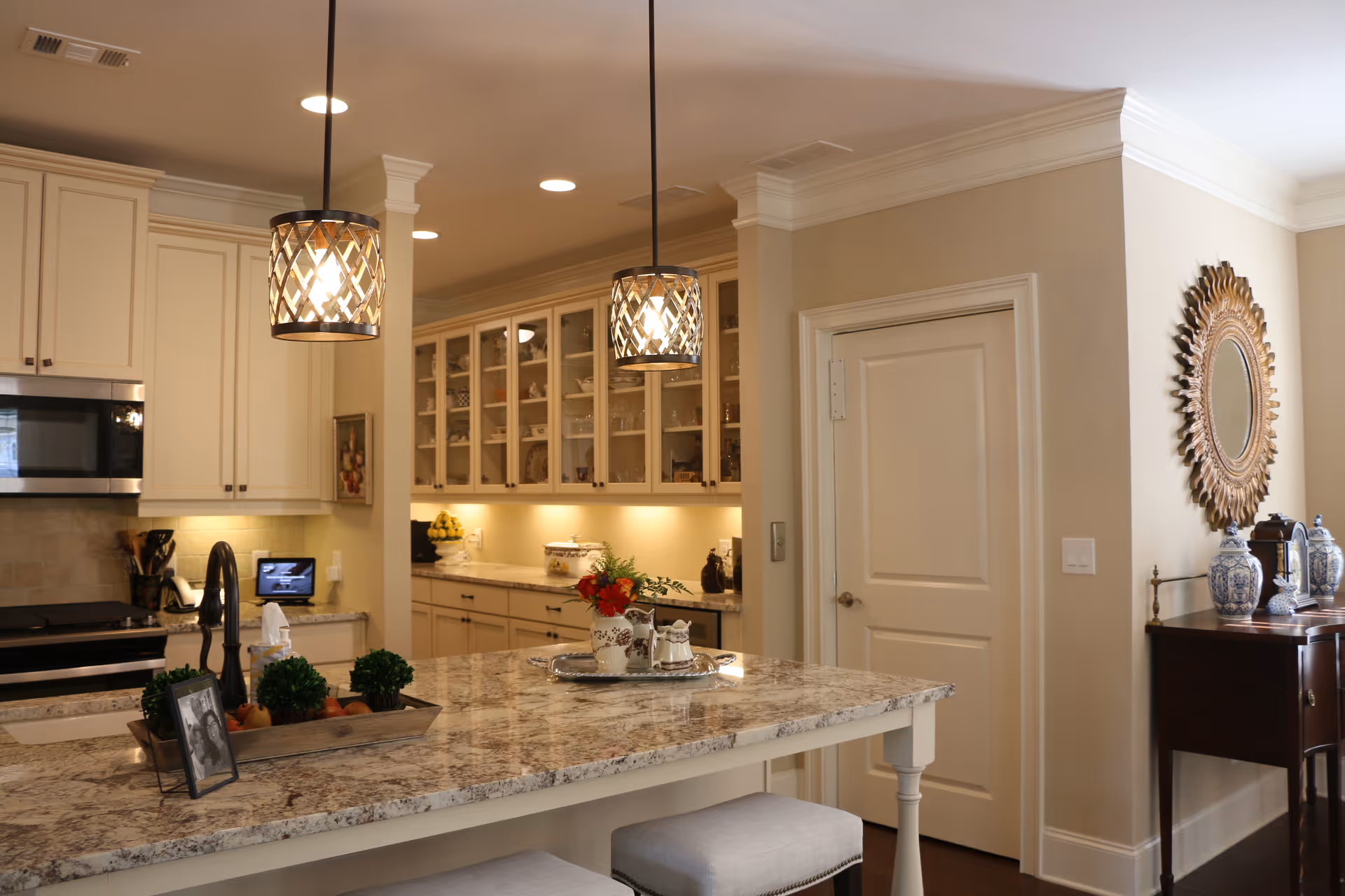 A bright and modern kitchen with granite countertops, white cabinetry, and two pendant lights hanging over a kitchen island with stools. The kitchen features a built-in microwave, a sink with a black faucet, and decorative items including a tray with plants and a framed photo on the island. There is a door on the right wall and a decorative mirror above a wooden side table with blue and white vases.