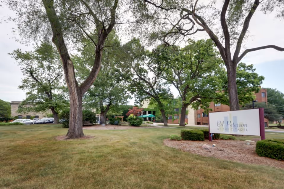 Outdoor view of PA Peterson at the Citadel facility showing a grassy area with large trees, a walking path, and a sign with the facility's name. The building is visible in the background with some parked cars on the left side.