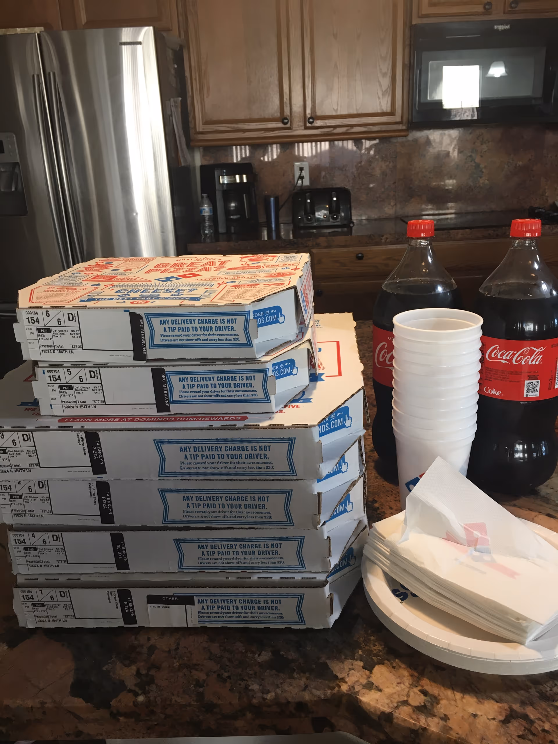 Stack of six Domino's pizza boxes on a kitchen counter with two bottles of Coca-Cola, a stack of white disposable cups, napkins, and plates. The kitchen has wooden cabinets, a stainless steel refrigerator, a coffee maker, a toaster, and a microwave.