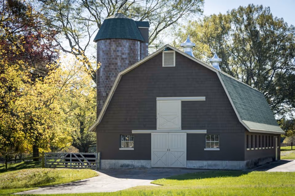 A large brown barn with a green roof and a cylindrical silo attached to its left side, surrounded by trees with autumn foliage and a grassy area in front.