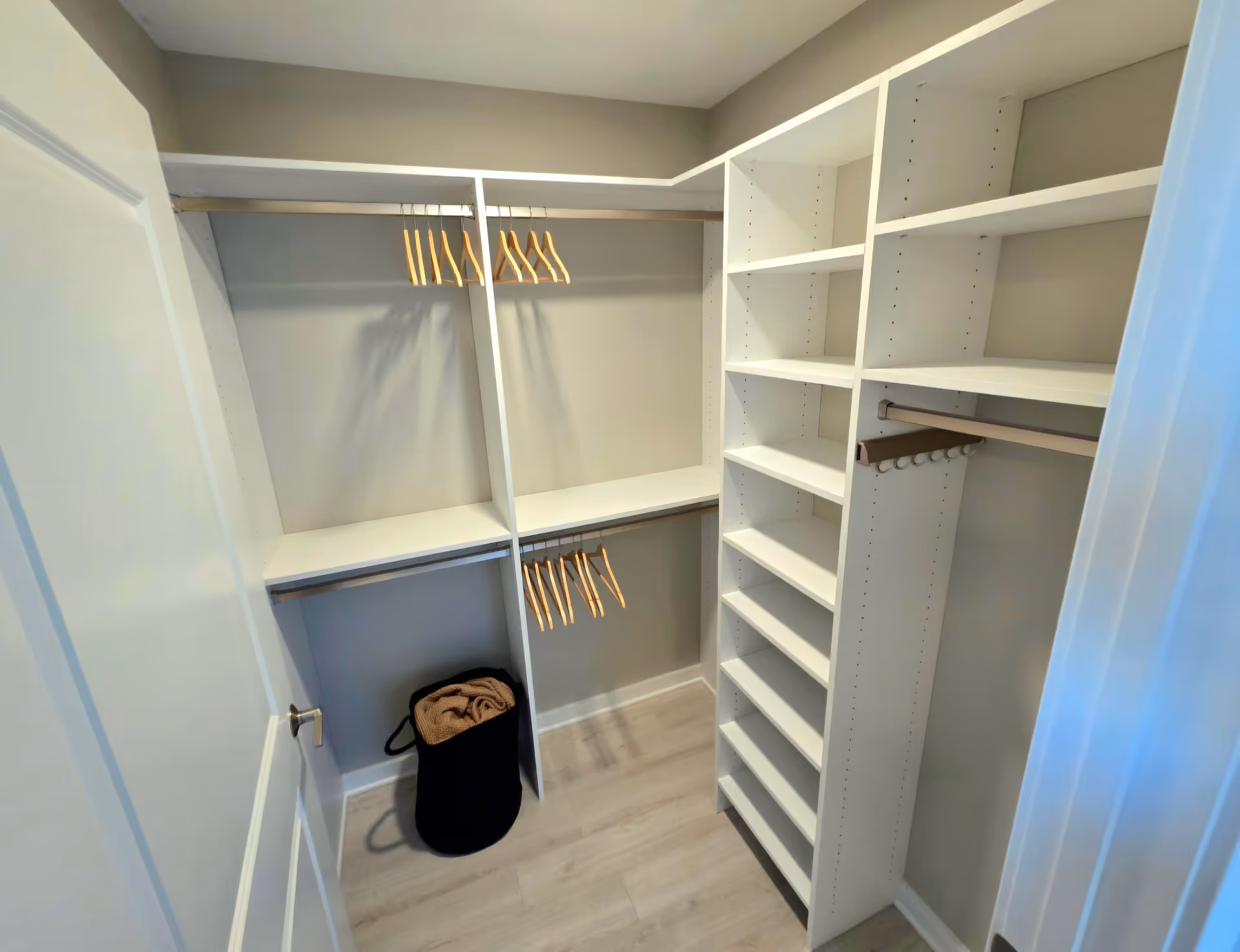 A walk-in closet with white shelving units and hanging rods. Several wooden hangers are hanging on the rods. There is a black basket on the floor containing a folded brown blanket or towel. The closet has light-colored wooden flooring and neutral-colored walls.