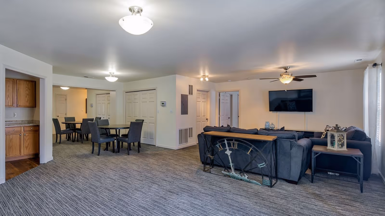 Interior view of a senior living facility common area featuring a living room with a large sectional sofa, a wall-mounted flat-screen TV, a ceiling fan with light, and a decorative clock piece behind the sofa. Adjacent to the living room is a dining area with two round tables and several chairs. The space has carpeted flooring, white walls, and multiple doors leading to other rooms.
