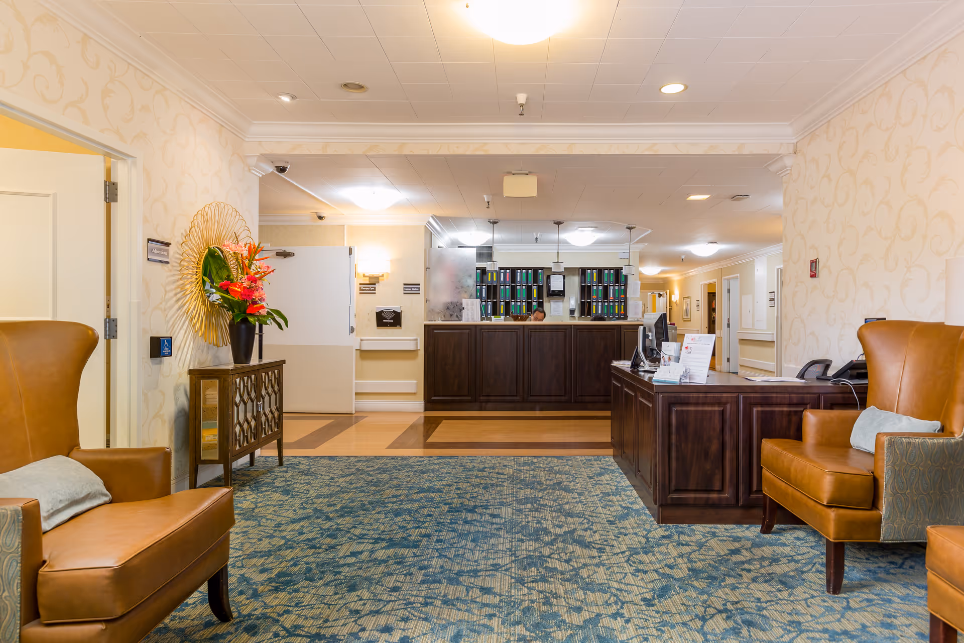 Reception area of Brentwood Health Care Center featuring a front desk with organized files behind it, two brown leather armchairs with cushions, a decorative console table with a vase of flowers, patterned wallpaper, and a blue patterned carpet.