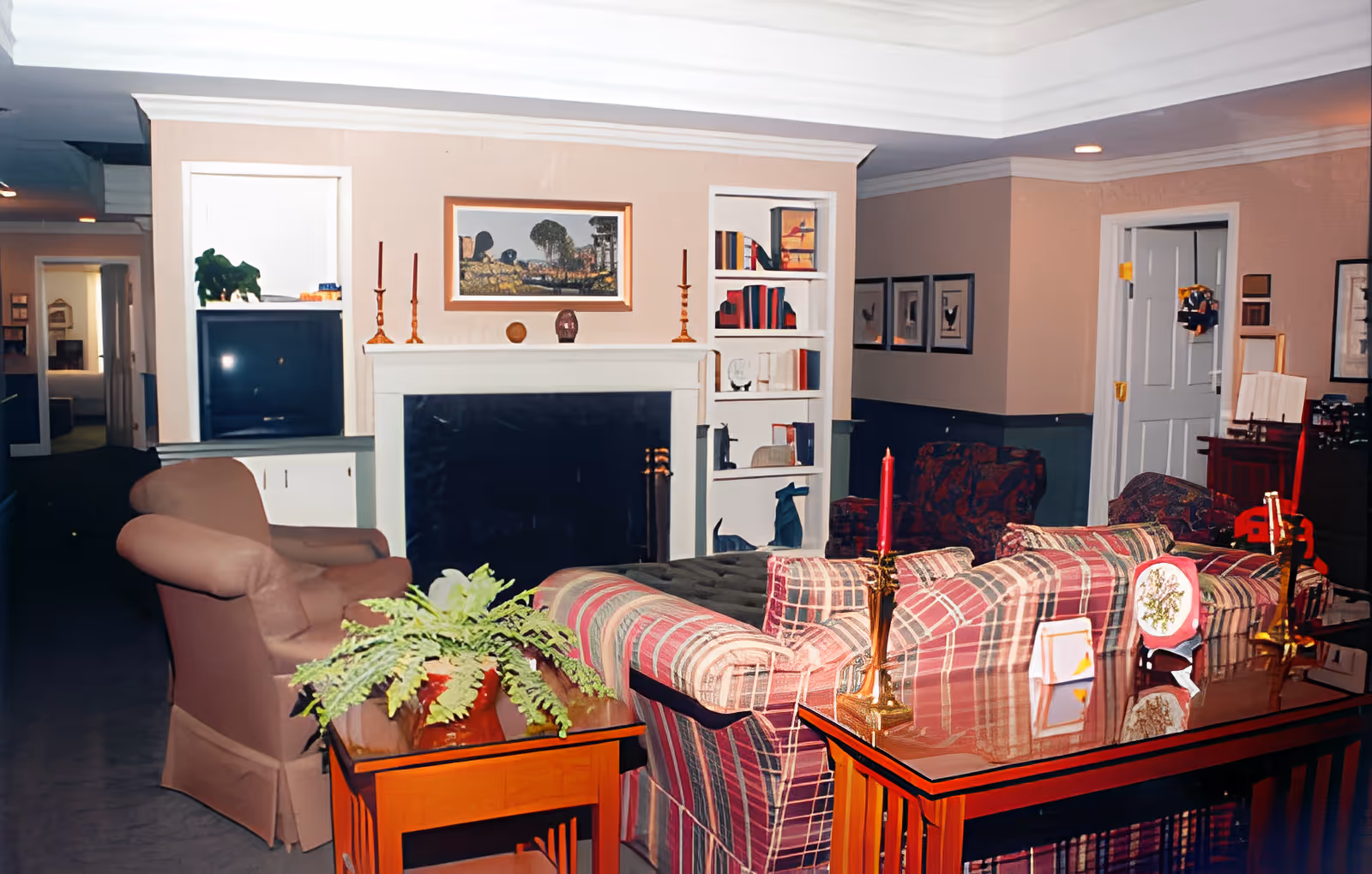 A cozy living room area in an assisted living facility featuring a plaid sofa, an armchair, a wooden coffee table with decorative items including a red candle and a small framed picture, a fireplace with candlesticks on the mantel, built-in shelves with books and decorations, and framed artwork on the walls.
