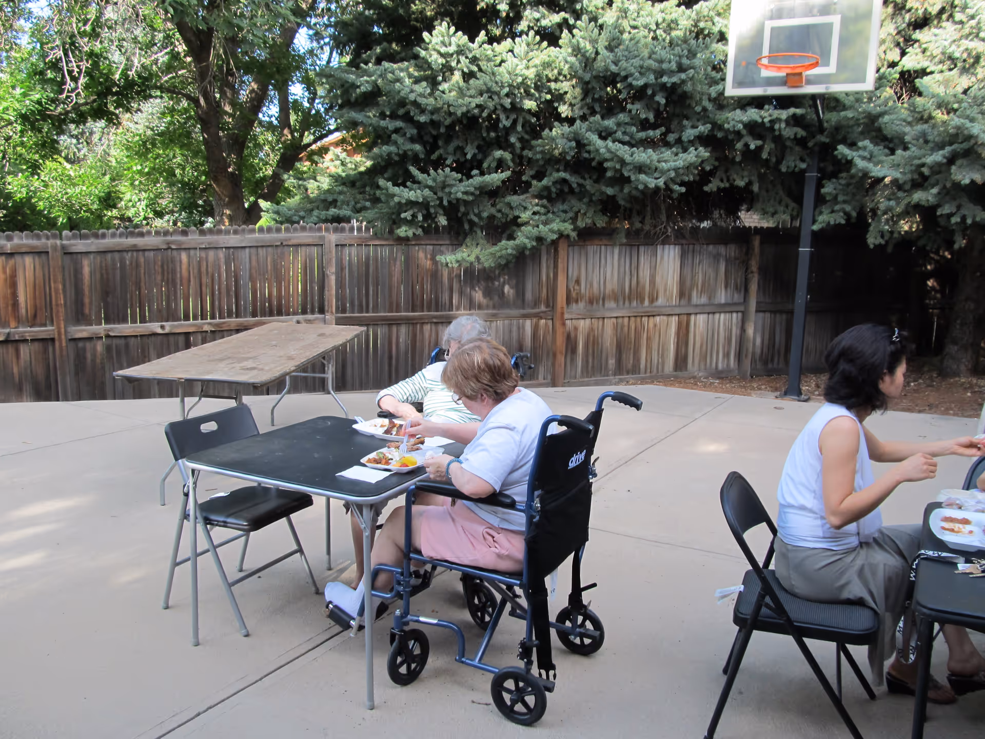 Three people sitting outdoors at folding tables eating a meal. Two elderly women, one in a wheelchair, and a younger woman are seated on a concrete patio with a wooden fence and trees in the background. A basketball hoop is visible behind them.