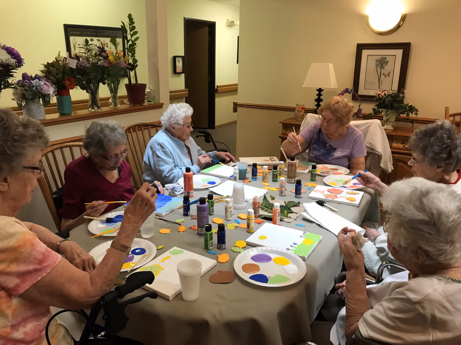 A group of elderly women seated around a table engaged in a painting activity with various paint bottles, brushes, and paper plates with paint on them. The room has a cozy interior with flowers on a shelf and framed artwork on the walls.