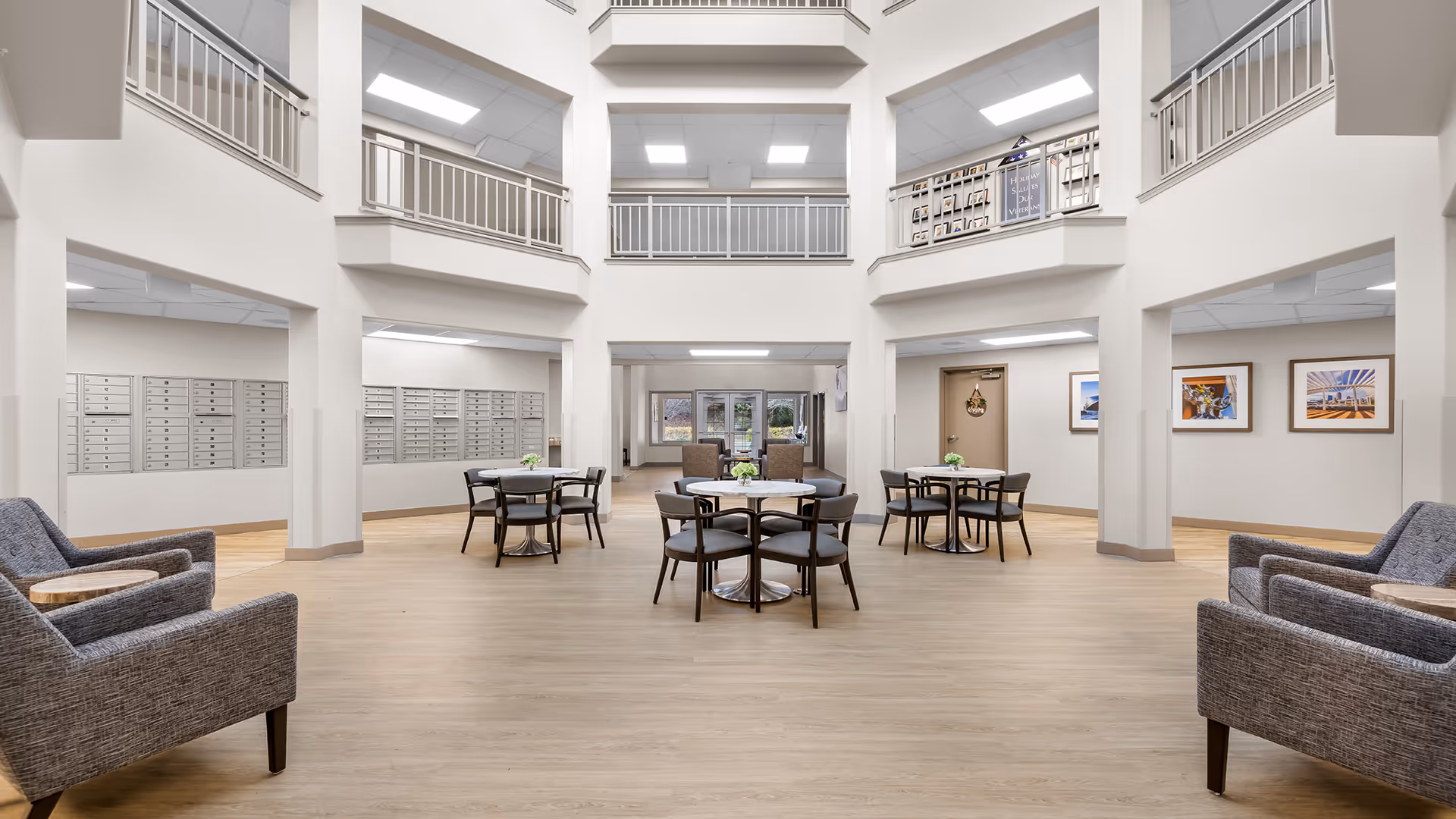Spacious and bright common area in a senior living facility with multiple round tables and chairs arranged in the center. Two gray upholstered armchairs with small wooden side tables are positioned on either side of the room. The area features light wood flooring, white walls, and a high ceiling with a second-floor balcony overlooking the space. Mailboxes line the walls on the left side, and framed artwork is displayed on the right wall. Large windows and glass doors at the far end allow natural light to enter.