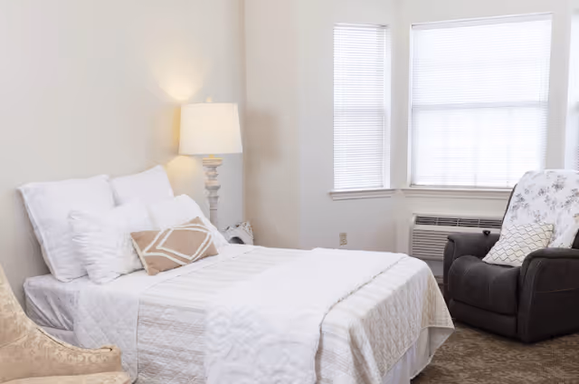 A cozy bedroom with a neatly made bed featuring white and beige bedding, a bedside table with a lamp, a cushioned armchair, and a large window with blinds letting in natural light.