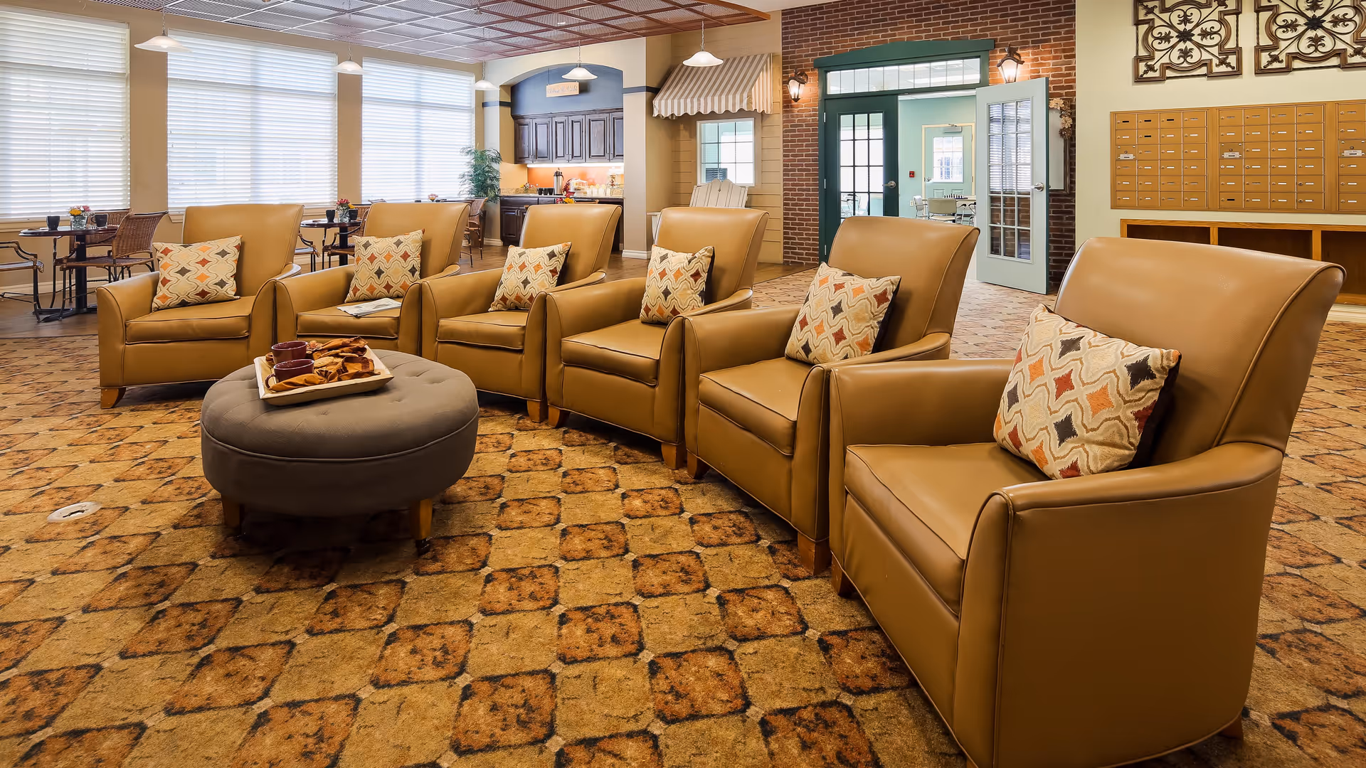 A cozy lounge area with five tan leather armchairs arranged in a row, each with a patterned cushion. In front of the chairs is a round gray ottoman with a tray of snacks. The room has a patterned carpet, large windows with blinds, a small kitchen area in the background, and a wall with mailboxes on the right side.