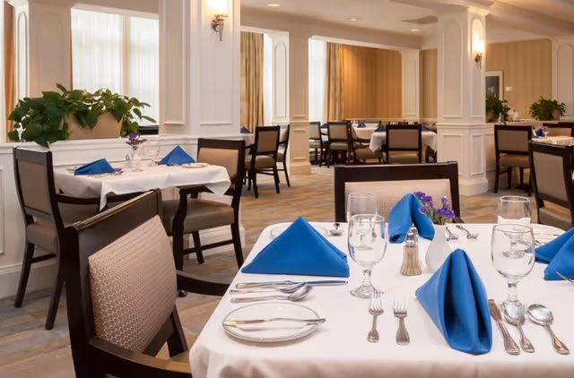 A well-lit dining room with tables covered in white tablecloths, each set with blue folded napkins, glassware, silverware, and small flower arrangements. The room features beige walls with white trim, large windows with curtains, and several chairs around each table.