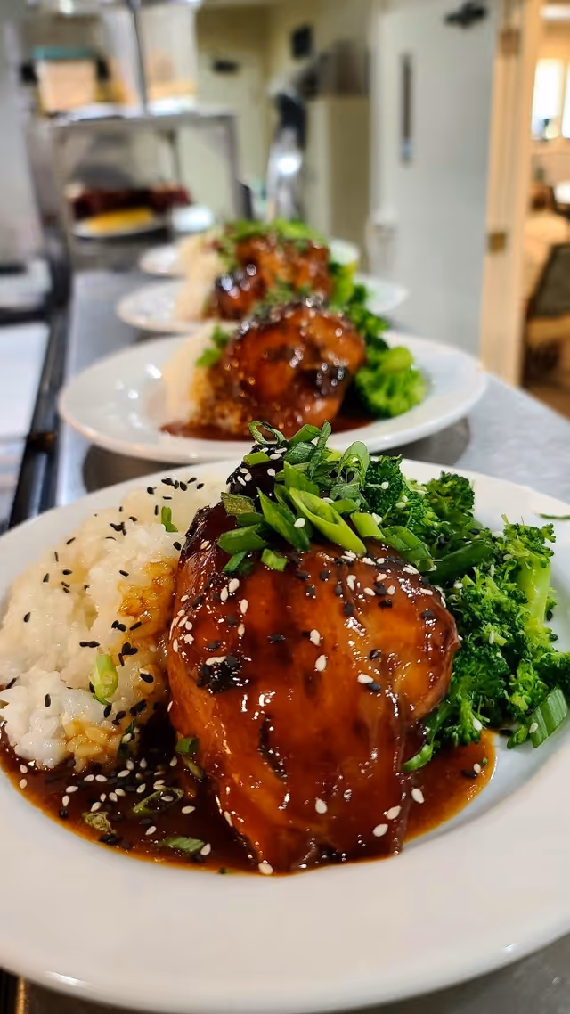 Close-up view of plates with a serving of glazed chicken topped with sesame seeds and chopped green onions, steamed broccoli, and white rice garnished with black sesame seeds, arranged in a row on a kitchen counter.