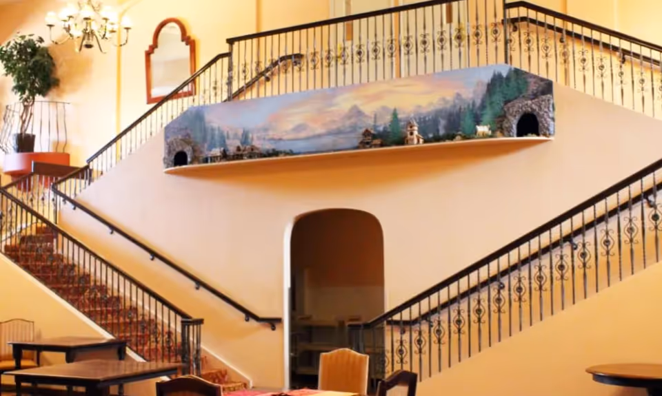Interior common area with a double staircase featuring decorative iron railings, dining tables, and a painted landscape mural above an arched doorway.