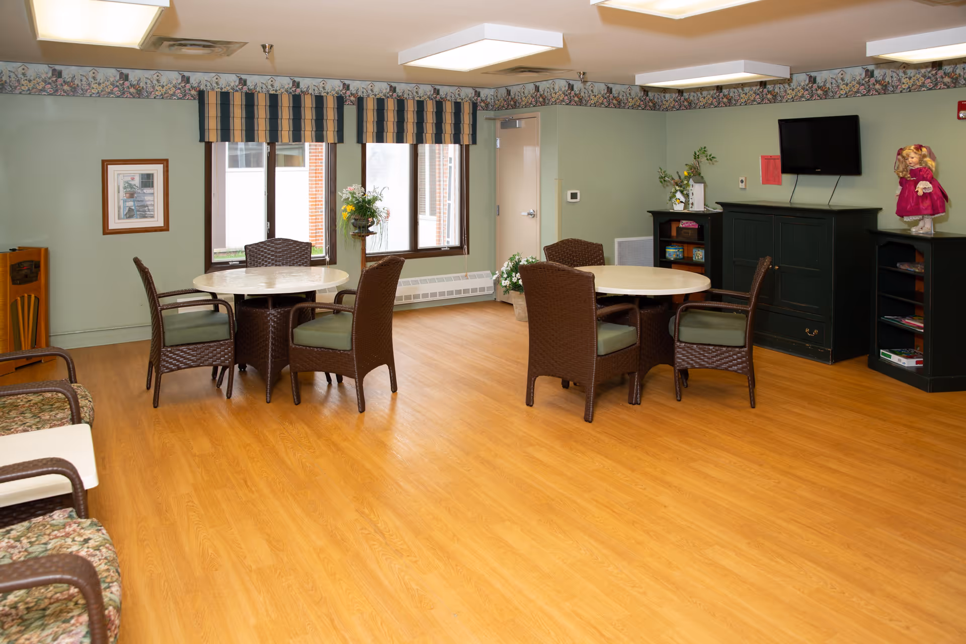 A common area with light green walls and wood flooring featuring two round tables each surrounded by four wicker chairs with green cushions. There are windows with striped valances, a TV mounted on a dark green cabinet, shelves with books and games, floral decorations, and a doll dressed in pink on one of the shelves.