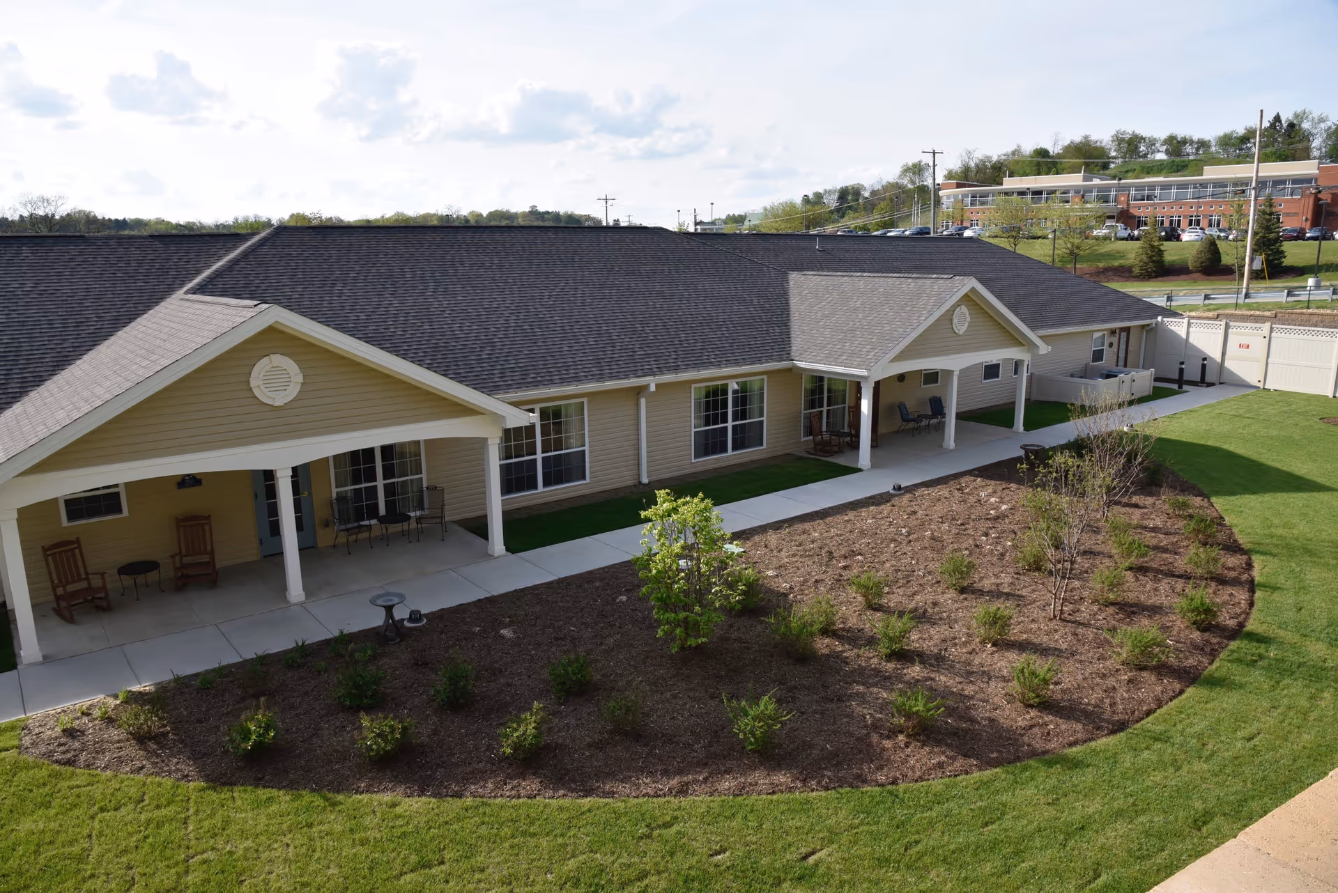 Exterior view of a single-story senior living facility building with beige siding and a dark shingled roof. The building features covered patios with rocking chairs and small tables. In front of the building is a landscaped garden area with small shrubs and a well-maintained lawn.