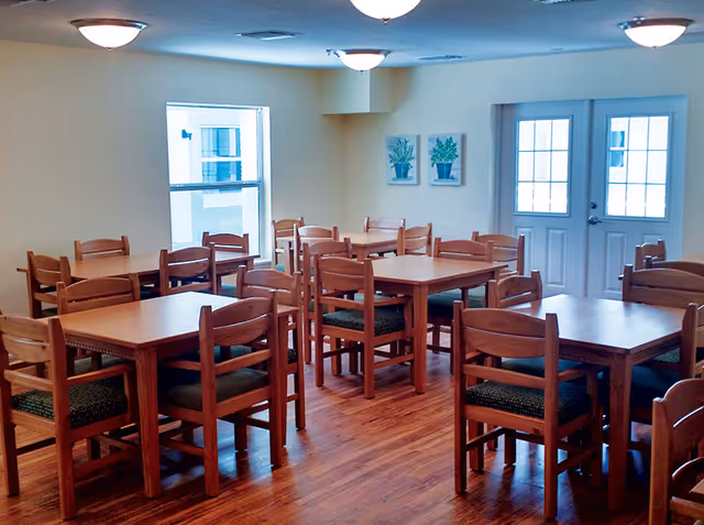 A dining room with multiple wooden tables and chairs arranged neatly on a wooden floor. The room has cream-colored walls, two framed plant pictures on the wall, a window, and a set of double doors with glass panes. Ceiling lights provide illumination.