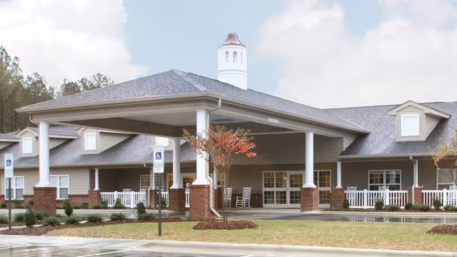 Front entrance of a senior care facility with a large covered porte-cochere, white columns, a porch with rocking chairs, and landscaped grounds.