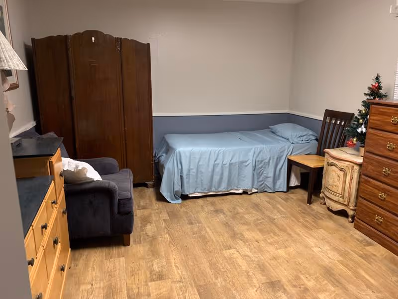 Small furnished bedroom with a single bed against the wall, an armchair, wooden dressers, a chair, and a folding room divider.