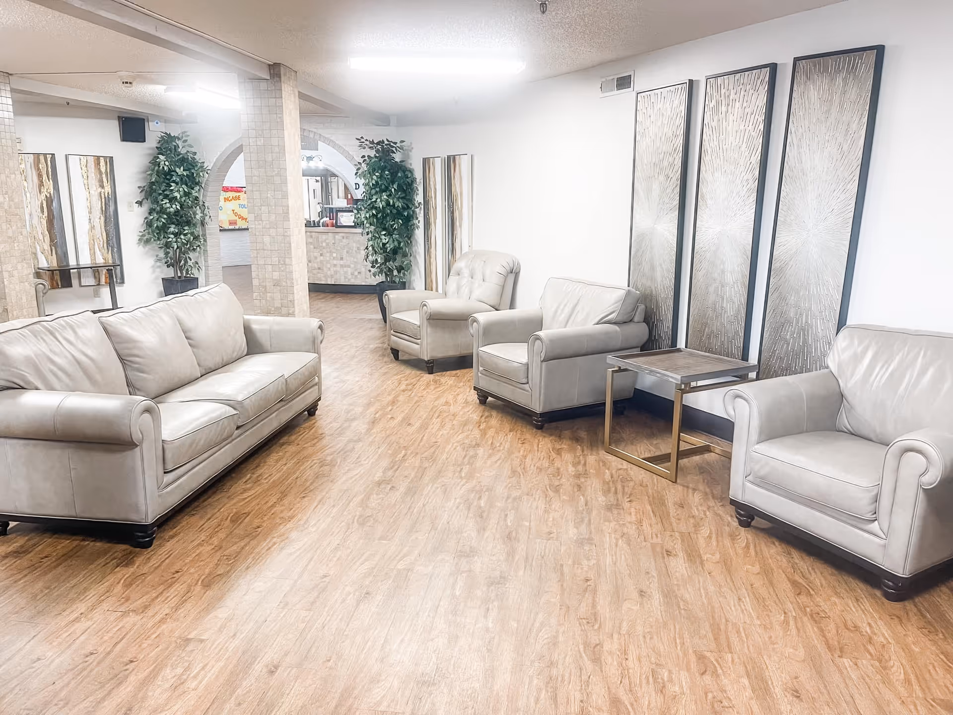 A bright and clean lounge area with light gray leather sofas and armchairs arranged on a wood floor. The walls are white and decorated with three large abstract art panels. There are two tall green plants near a tiled column and an arched opening leading to a reception desk in the background.