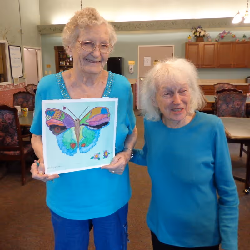 Two elderly women standing inside a room with tables and chairs. One woman is holding a colorful painting of a butterfly. Both women are smiling and wearing blue tops. The room has wooden cabinets, floral decorations, and soft lighting.