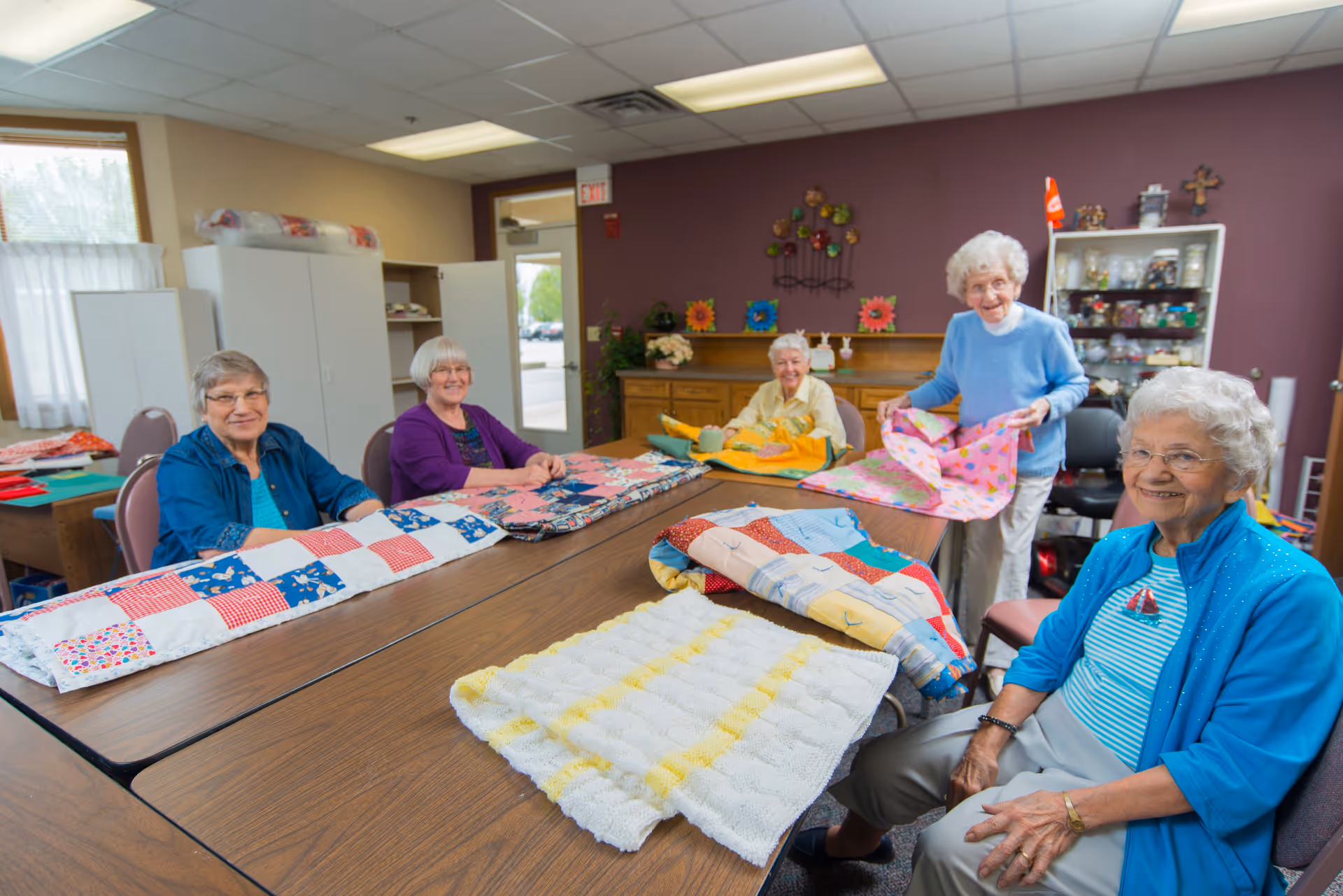 Five elderly women sitting and standing around a large table in a craft room, smiling and displaying colorful handmade quilts. The room has purple walls, cabinets, and shelves with various craft supplies and decorations.