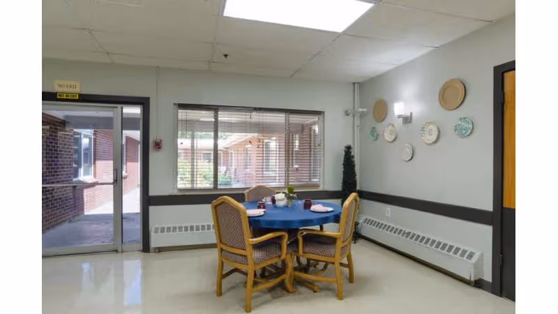 A small dining area inside a facility with a round table covered by a blue tablecloth and four wooden chairs with cushioned seats. The table is set with plates, cups, and a small centerpiece. There is a large window with blinds showing an outdoor brick courtyard. The walls are light-colored with decorative plates hanging on one side. A glass door labeled 'No Exit' leads outside.