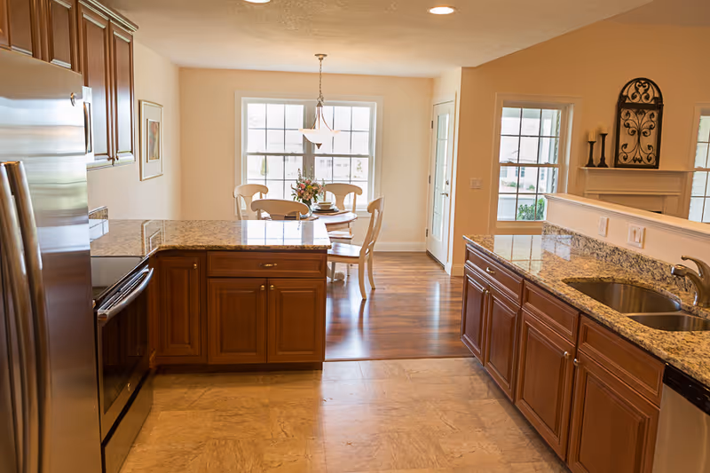 A bright kitchen with wooden cabinets, granite countertops, a stainless steel refrigerator, oven, and a double sink. In the background, there is a dining area with a round wooden table, four chairs, a hanging light fixture, and a window letting in natural light.