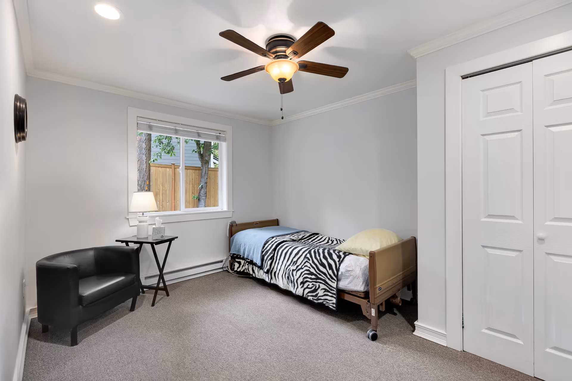 A simple senior care bedroom with a single hospital-style bed covered with a zebra-patterned blanket and a yellow pillow. The room has light gray walls, a ceiling fan with a light, a window showing a wooden fence and trees outside, a small black armchair, and a small table with a lamp and tissue box. There is a white double-door closet on the right side.