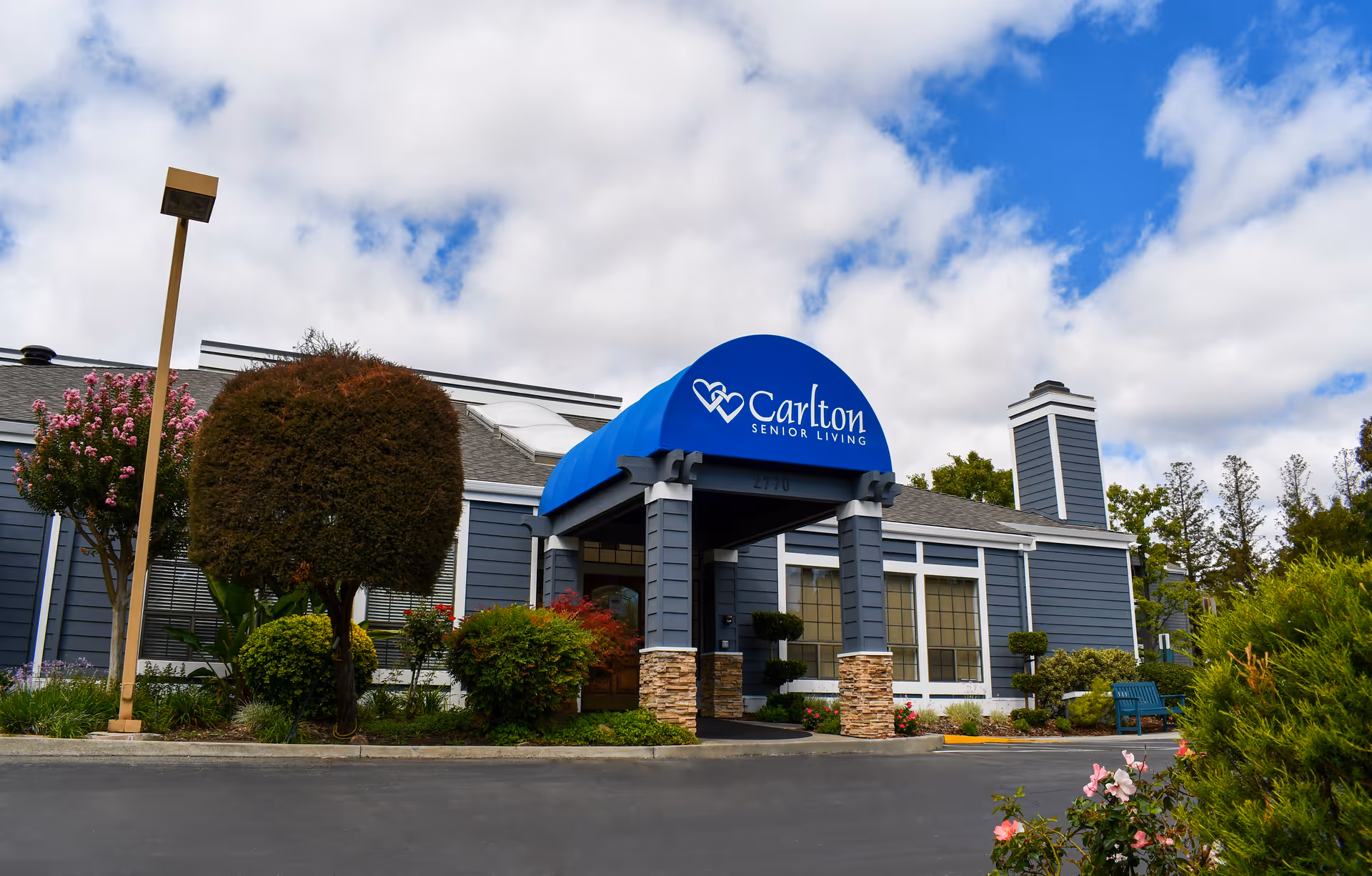 Front entrance of a Carlton Senior Living building with a blue awning, landscaped shrubs, and a cloudy sky.