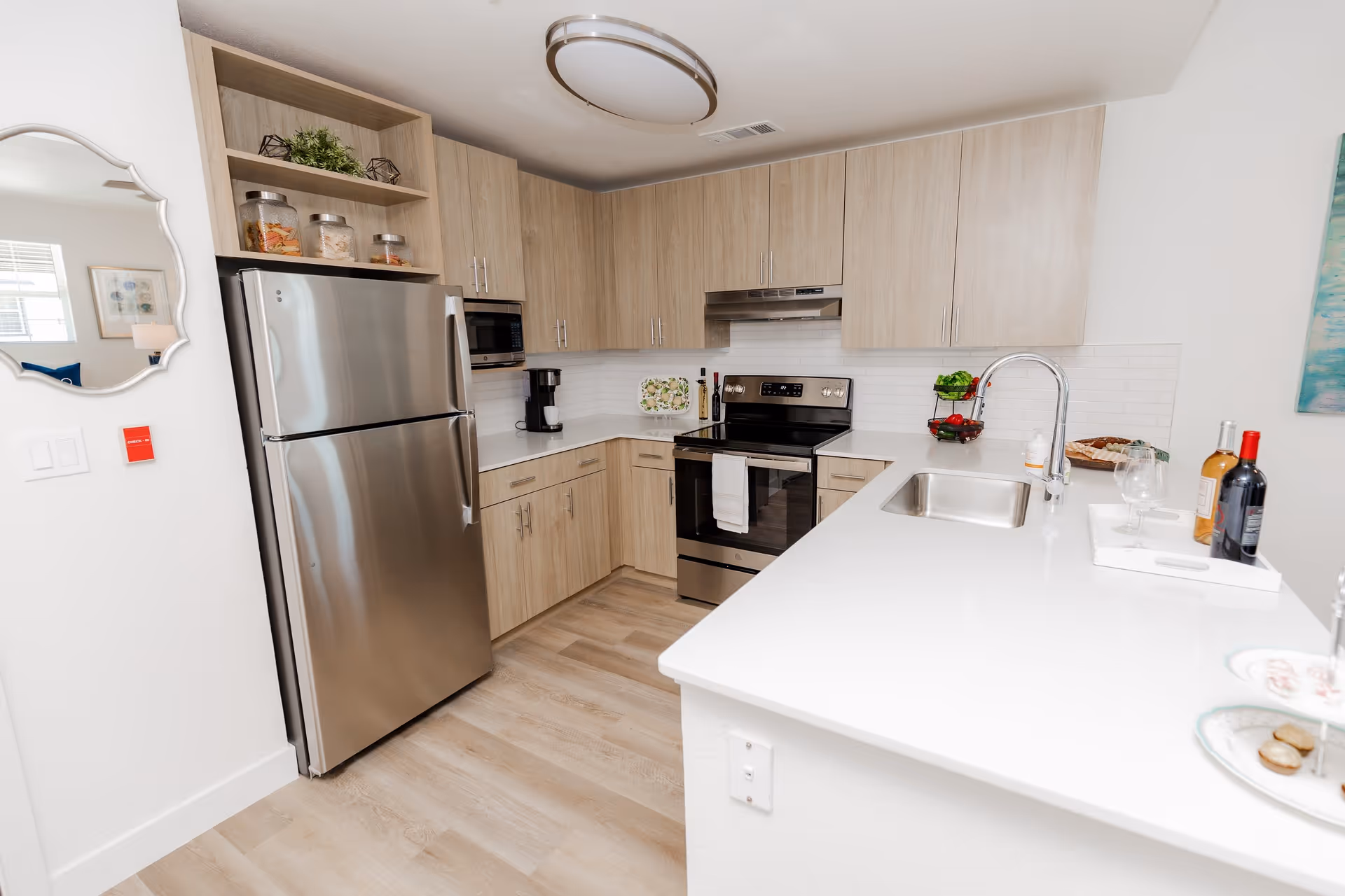 Modern kitchen with light wood cabinets, stainless steel refrigerator and oven, white countertops, a sink with a curved faucet, and various kitchen items including a coffee maker, wine bottles, and a fruit basket.