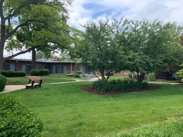 A well-maintained outdoor garden area at Delmar Gardens of Creve Coeur featuring green grass, trimmed bushes, several trees, a wooden bench, and a single-story brick building in the background under a partly cloudy sky.