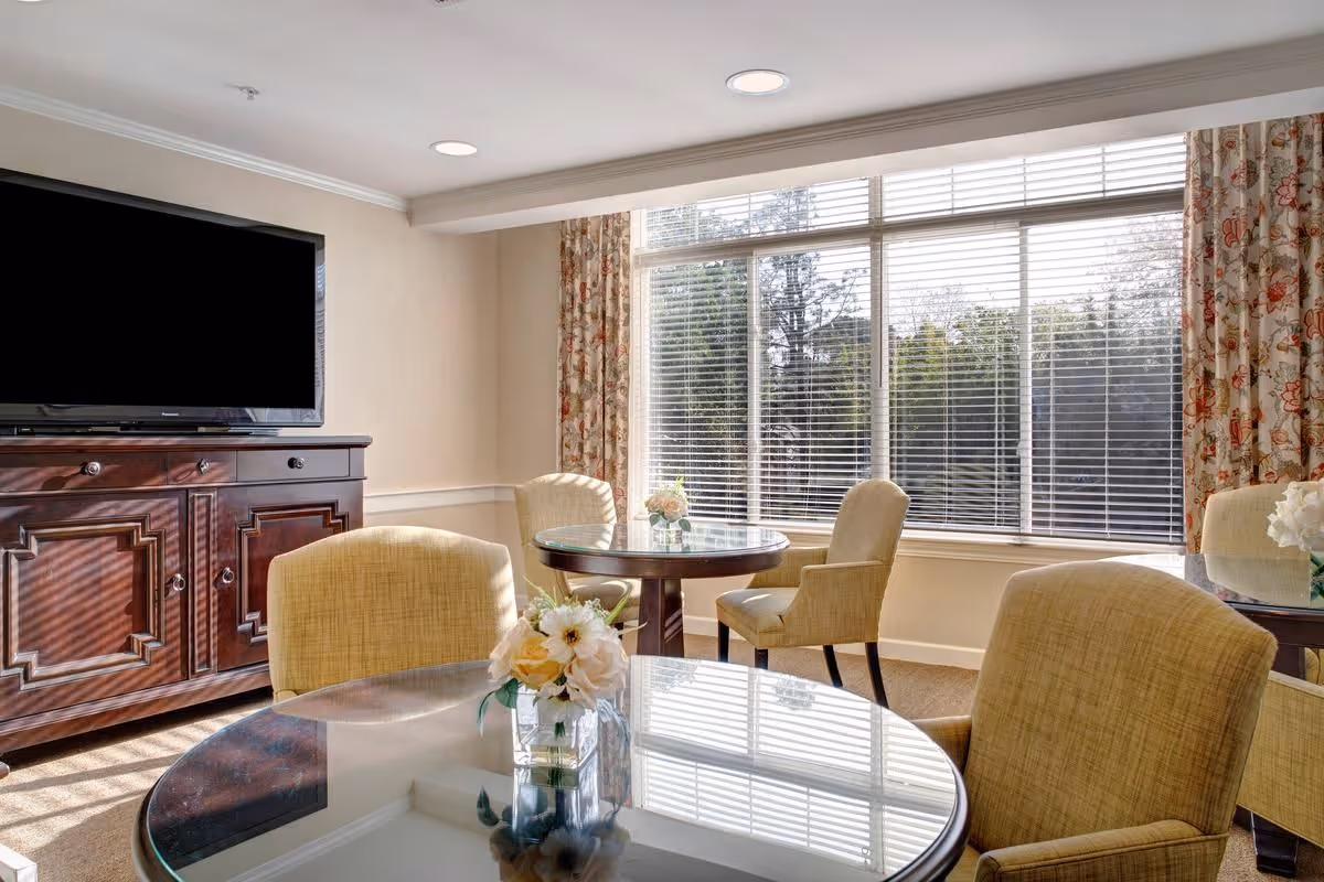 Sunlit common dining area with round glass-top tables, upholstered chairs, a wooden TV cabinet and large windows with blinds.