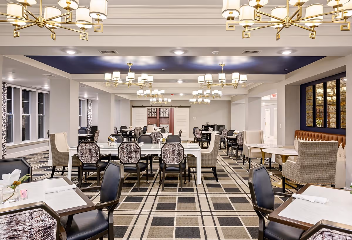 Spacious, well-lit dining room with multiple tables and chairs, chandeliers, and patterned carpeting in a senior living facility.