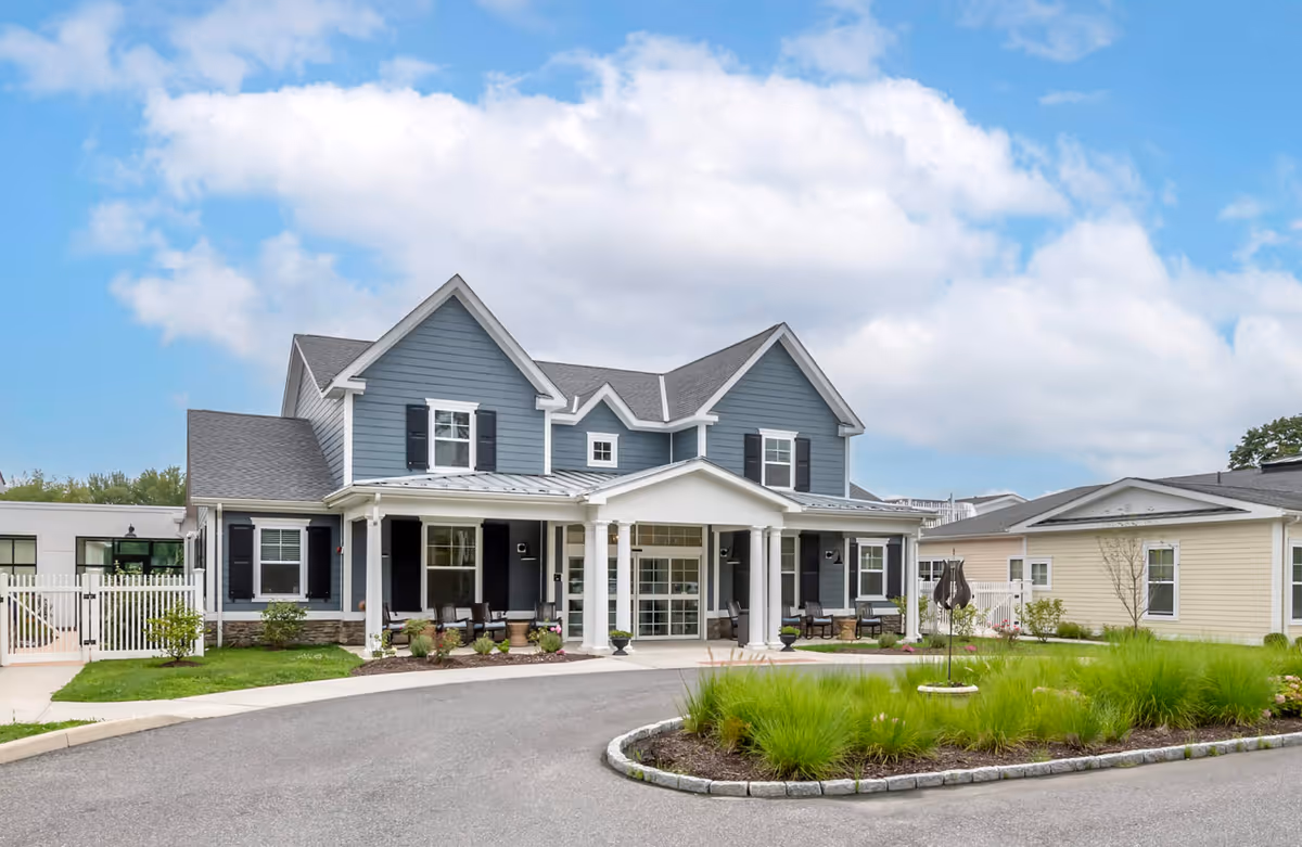 Front exterior view of a two-story senior living facility building with blue siding, white trim, and black shutters. The entrance features a covered porch with white columns and seating. There is a circular driveway with landscaped greenery in front of the building under a partly cloudy sky.