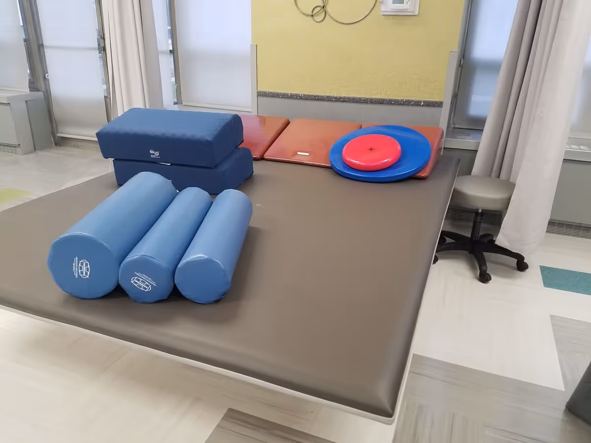 A therapy or rehabilitation room with a large padded table holding various physical therapy equipment including blue cylindrical bolsters, rectangular cushions, and circular balance discs. The room has light-colored flooring, windows with blinds, and a stool nearby.