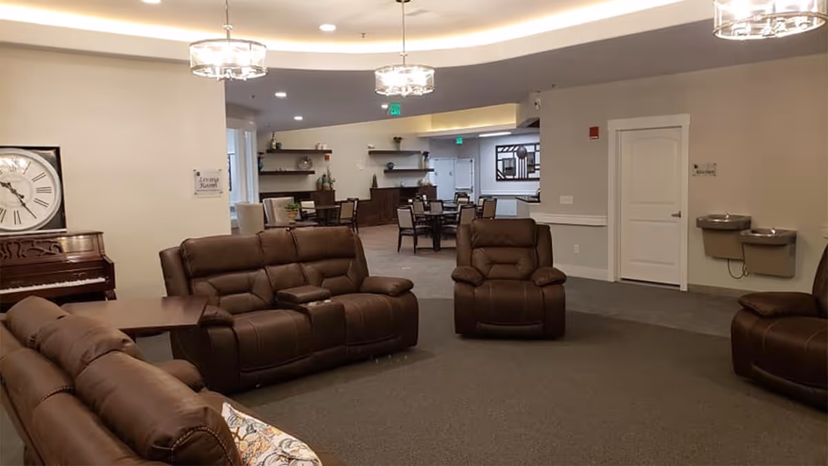 A spacious assisted living common area with brown leather recliner sofas and chairs arranged around a carpeted floor. In the background, there are tables and chairs for dining or activities, a piano with a large clock on the wall above it, and two water fountains mounted on the wall near a closed white door. The room is well-lit with ceiling lights and has neutral-colored walls.