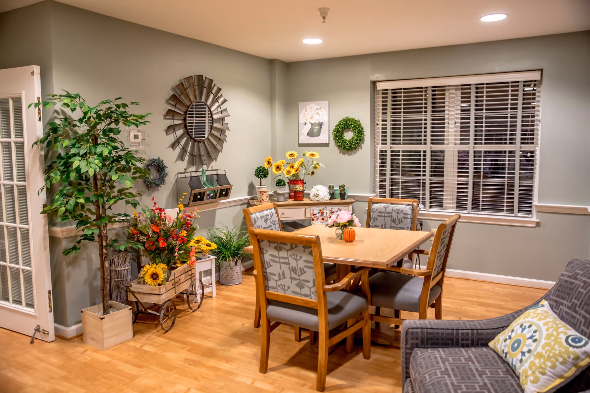 A cozy dining area in an assisted living facility featuring a wooden table with four cushioned chairs, a small side table with decorative plants and flowers, a large potted plant, a decorative mirror on the wall, and a window with white blinds. The room has light green walls and wooden flooring.