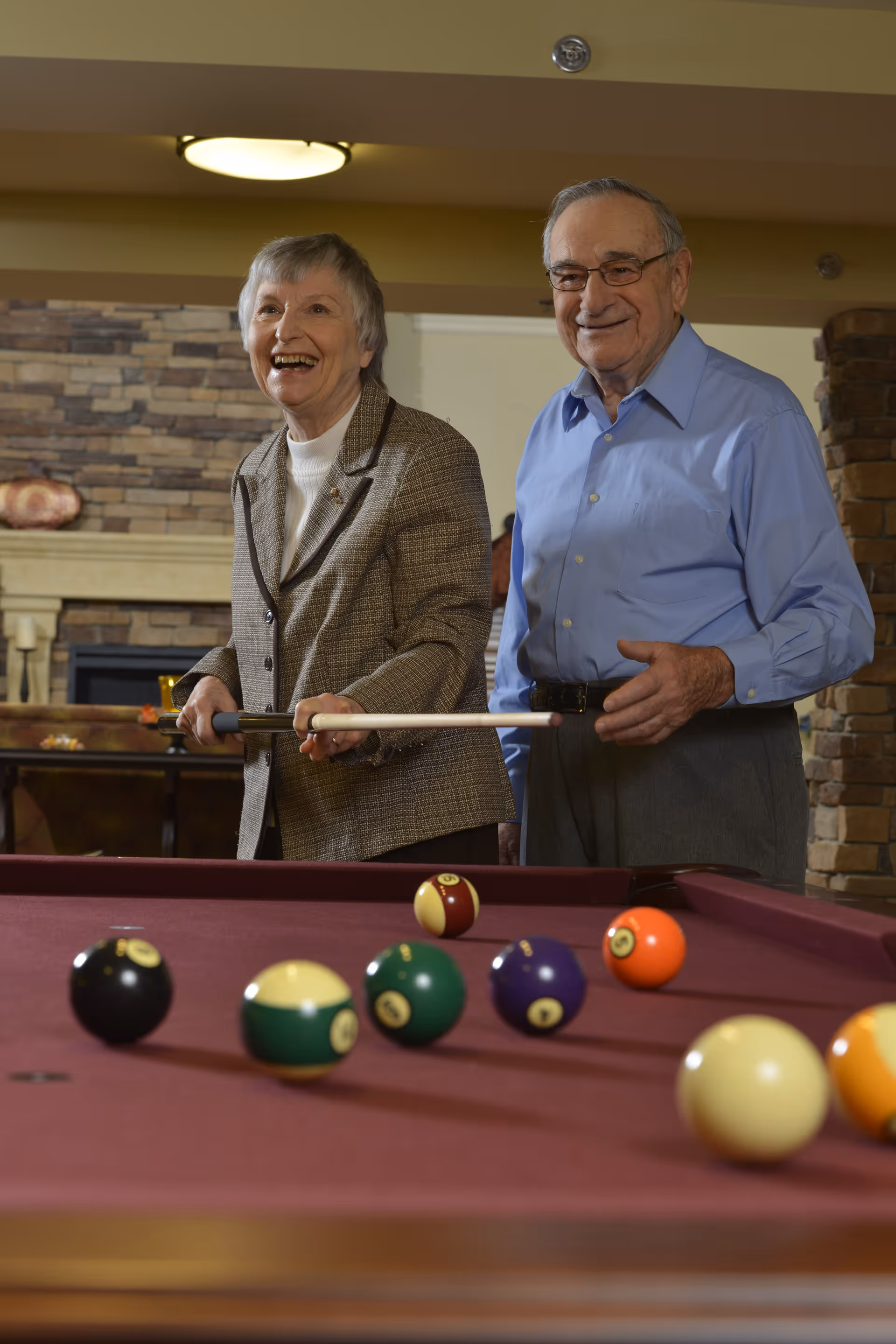 An elderly woman and man smiling and playing pool together in a cozy room with a stone fireplace and warm lighting.