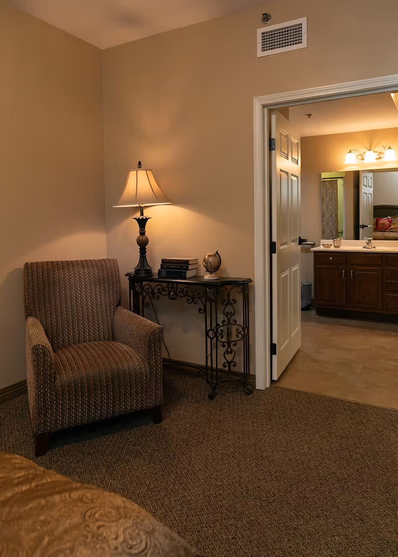 A cozy corner of a senior living facility room featuring a patterned armchair next to a decorative metal console table with a lamp, books, and a small globe. An open door reveals a bathroom with a large mirror, wooden cabinets, and a sink.