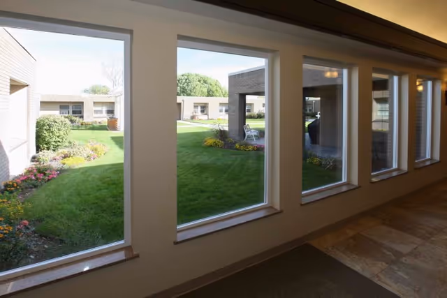 View through a row of large windows showing a well-maintained outdoor courtyard with green grass, flower beds, and a covered seating area in a senior living facility.