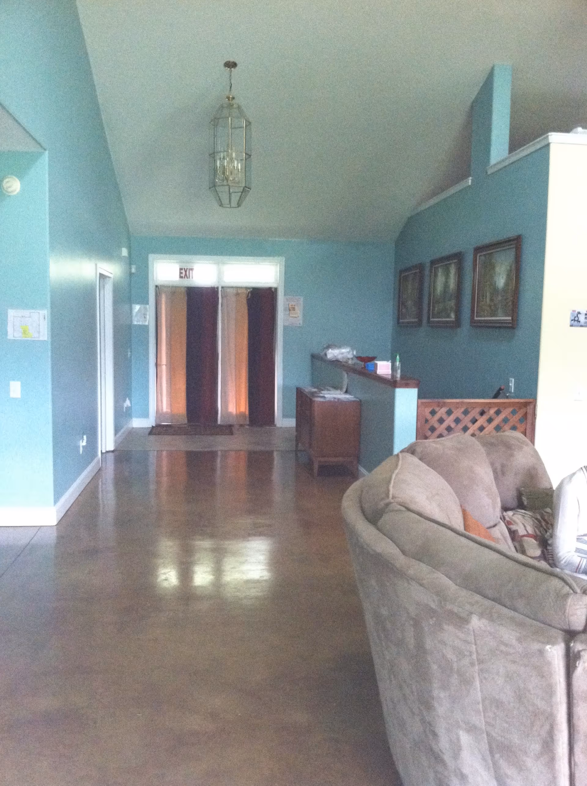 Interior view of a living room area with light blue walls, a brown sectional sofa on the right, three framed pictures on the wall, a small wooden cabinet, and an exit door with curtains at the far end. The floor is polished concrete and a hanging light fixture is visible on the ceiling.