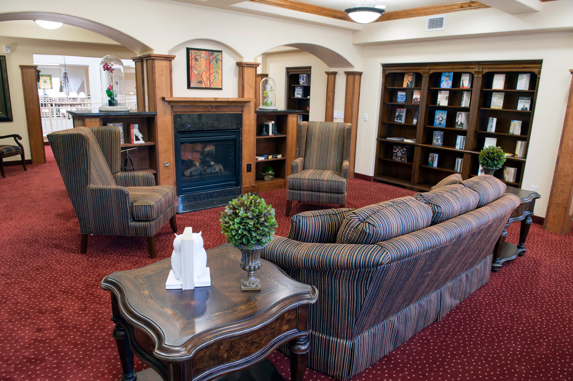 A cozy living room area with a striped sofa and two matching armchairs arranged around a wooden coffee table with a small green plant and white decorative items. The room features a fireplace with wooden built-in shelves on either side, a large bookshelf filled with books, and a red carpet with a subtle pattern. The walls are light-colored with wooden trim and arches.