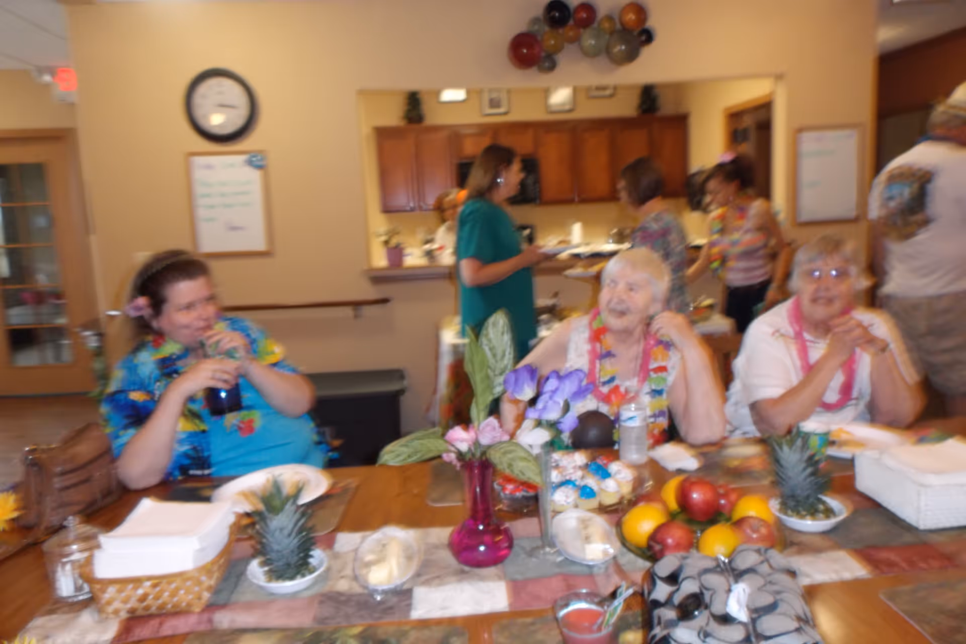 Several older adults and staff socializing around a decorated dining table with fruit, flowers, and plates in a communal dining area.