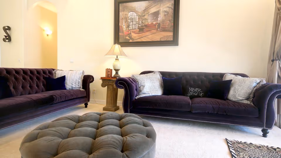 A living room with two dark purple tufted sofas facing each other, a large tufted gray ottoman in the center, a wooden side table with a lamp, and a framed painting on the wall. The room has light-colored walls and carpet, with curtains on the right side.