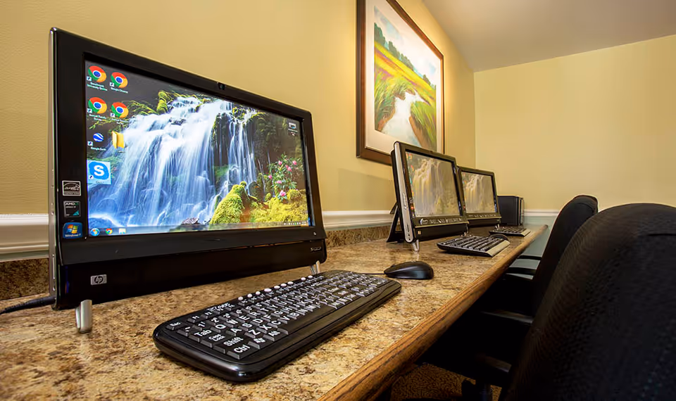 A row of desktop computers with keyboards and mice on a long granite countertop in a room with beige walls. There are three black office chairs in front of the computers, and a framed landscape painting hangs on the wall above the computers.