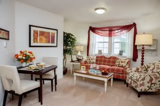 A cozy living room with a plaid sofa, floral armchair, white coffee table, and a small dining table with two chairs. The room is decorated with a red sheer curtain, a floor lamp, a table lamp, a potted plant, and framed artwork on the wall.
