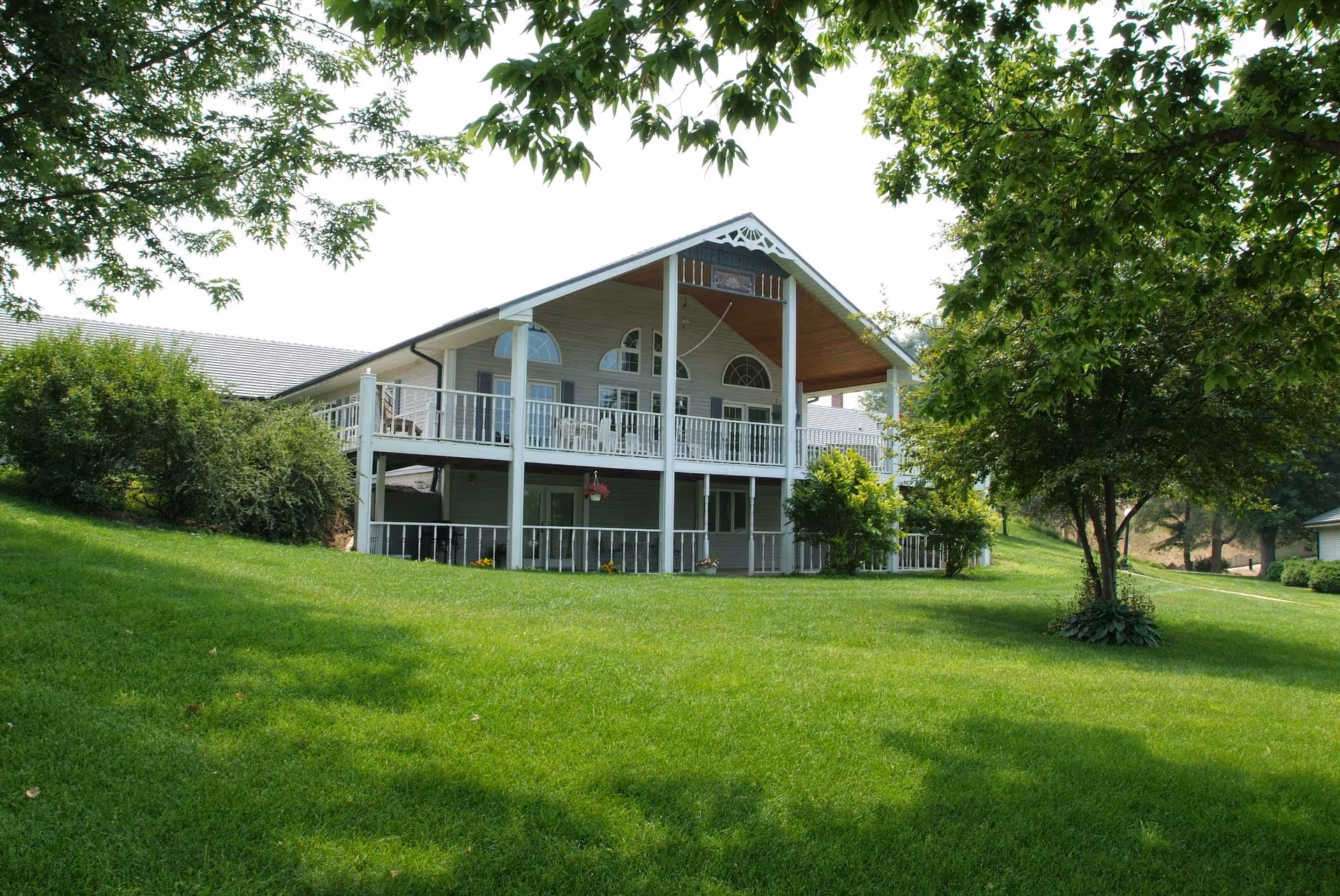 A two-story building with a large covered balcony on the upper floor and a patio area below, surrounded by green grass, trees, and bushes under a bright sky.