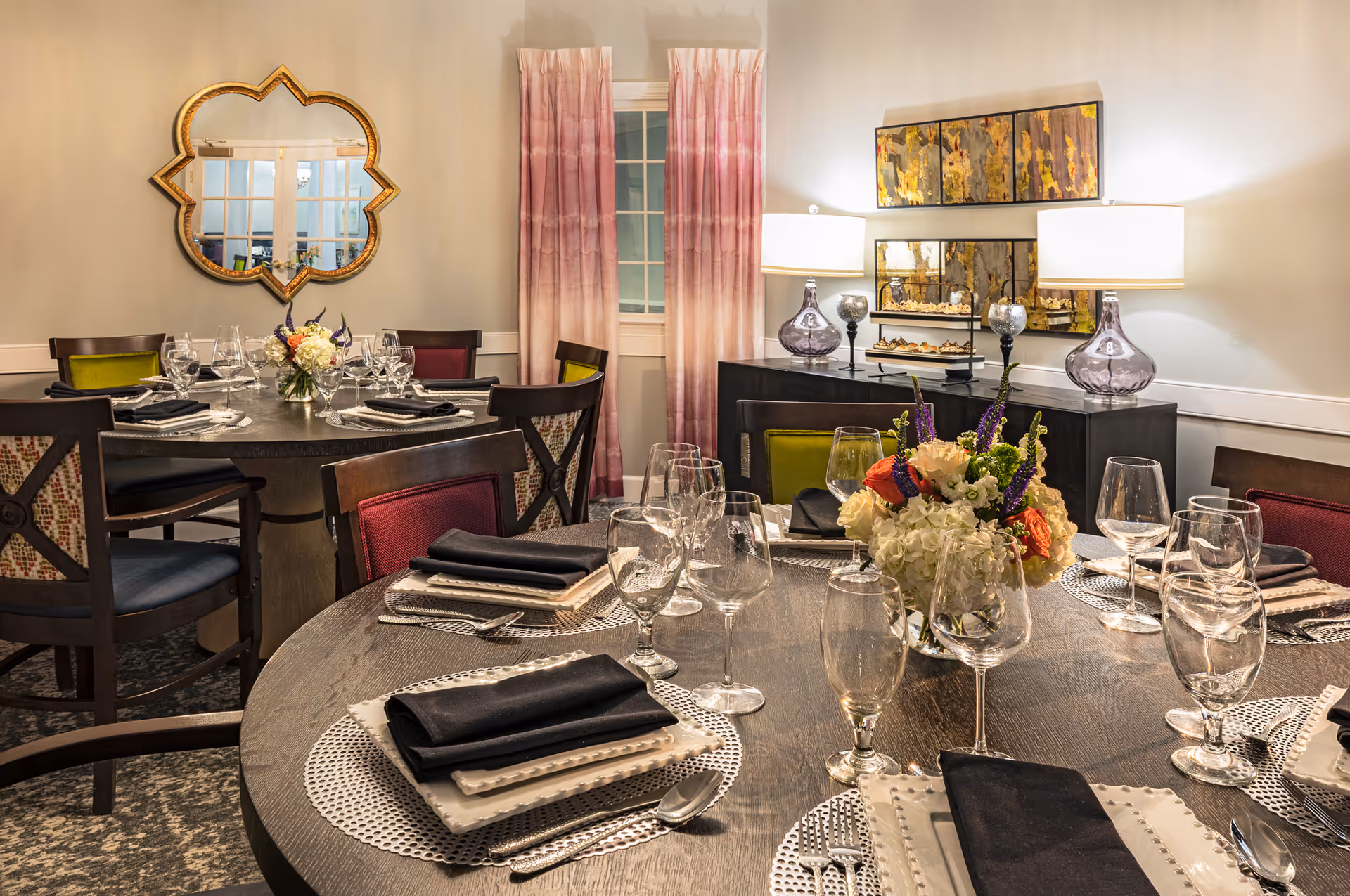 Dining room with round tables set with plates, black napkins, glassware and floral centerpieces, with decorative mirror, lamps and artwork in the background.