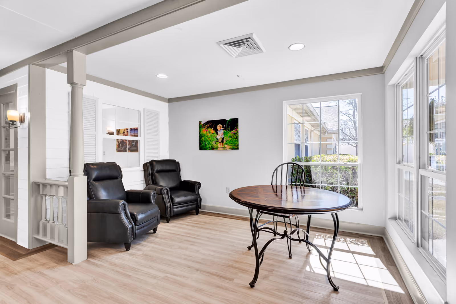 A bright room with two black leather armchairs positioned against a white wall, a round wooden table with a black metal frame, and large windows letting in natural light. A colorful painting of a child in a garden hangs on the wall.